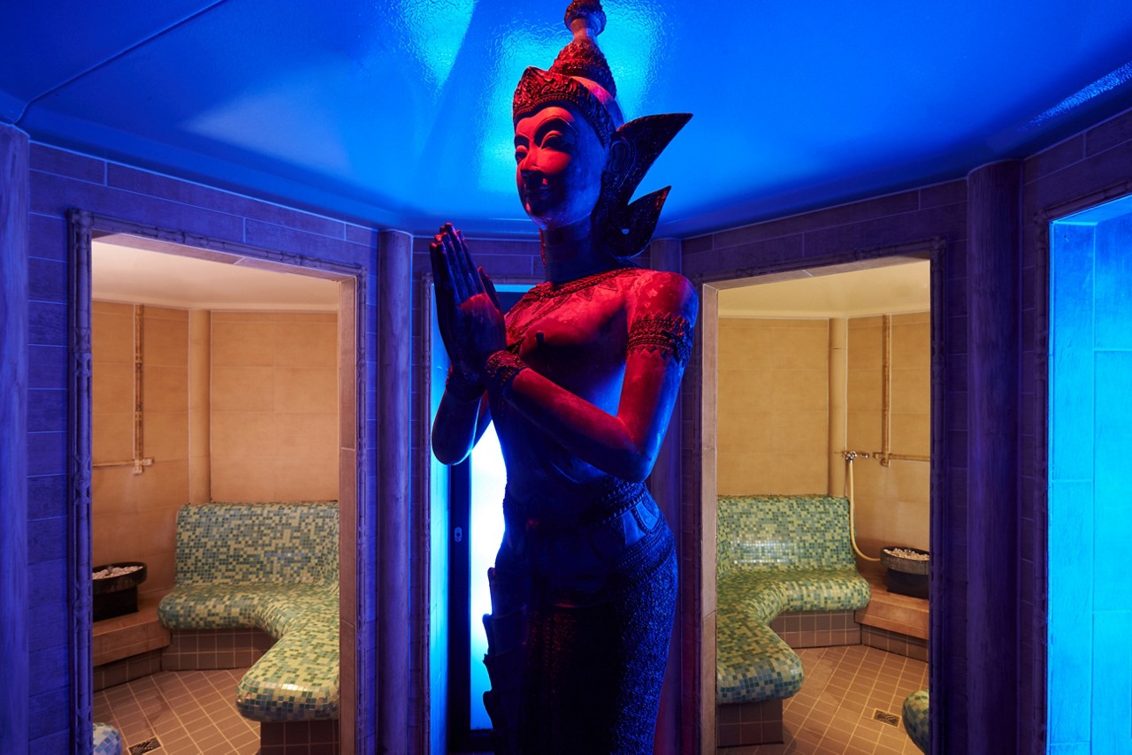 Statue with hands pressed together stands centered, illuminated blue-red, between two open tiled steam rooms. Mosaic benches curve along walls; small stone heaters sit in corners under warm beige lighting.